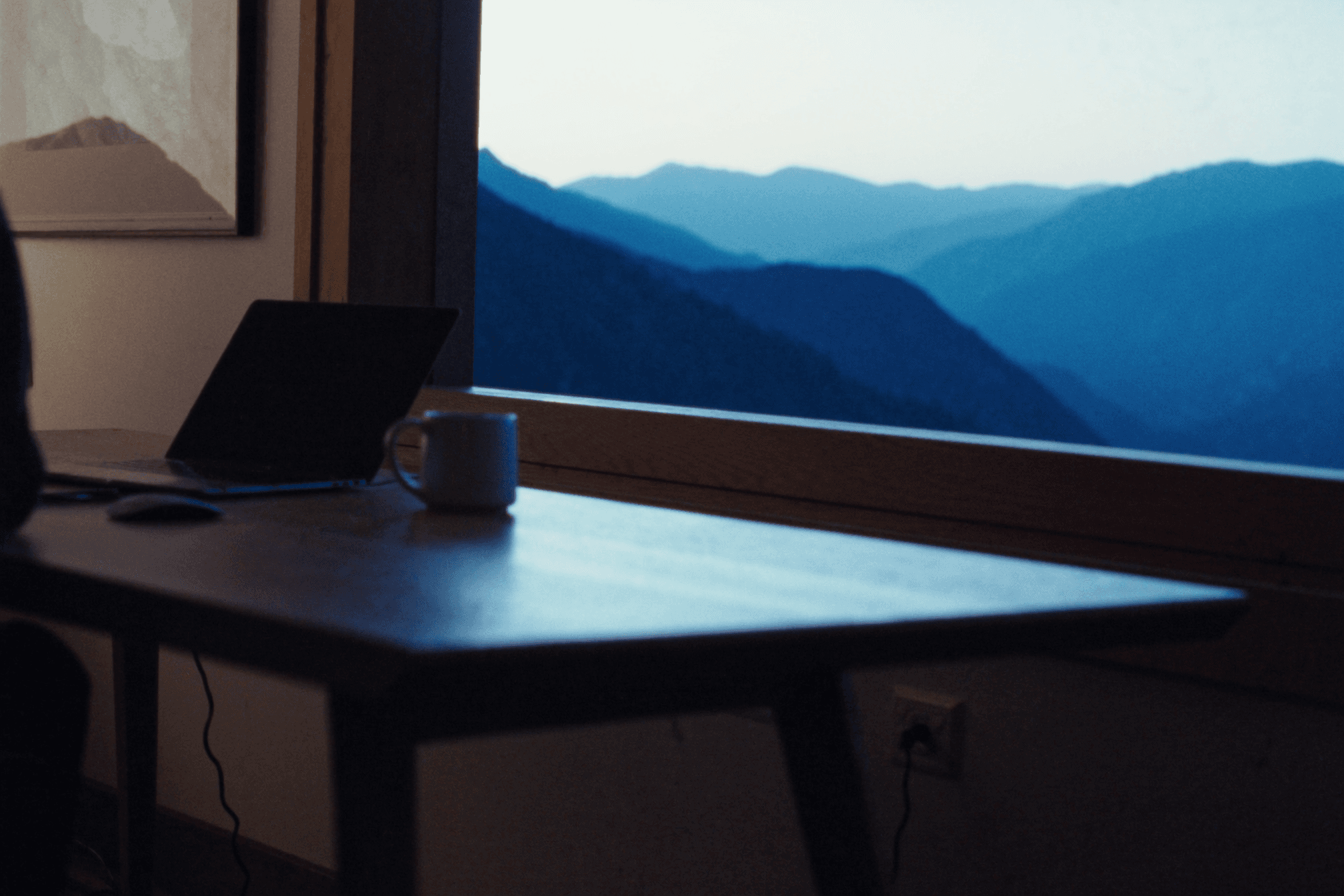 A desk with a laptop and coffee mug by a window overlooking misty mountains