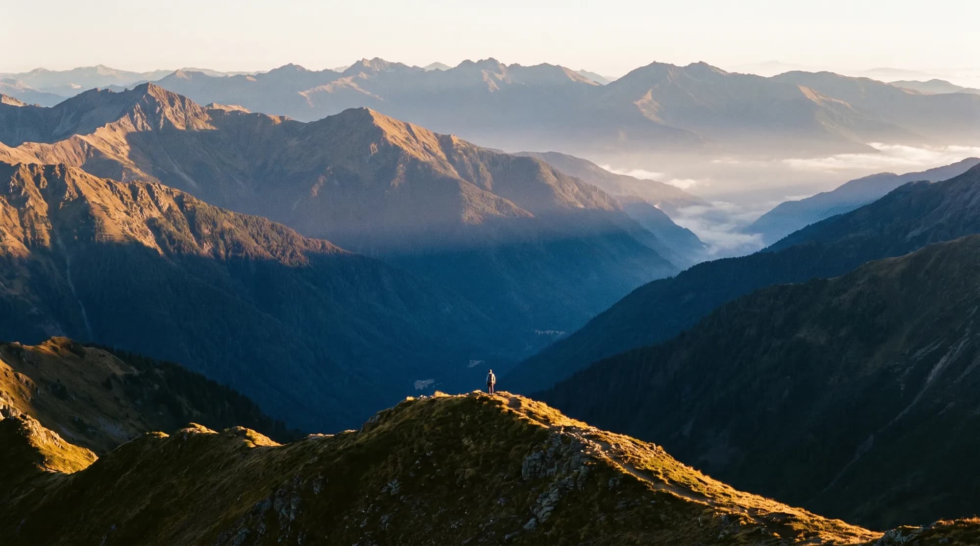 A lone hiker on a high ridgeline at sunrise, with layered mountain ranges behind